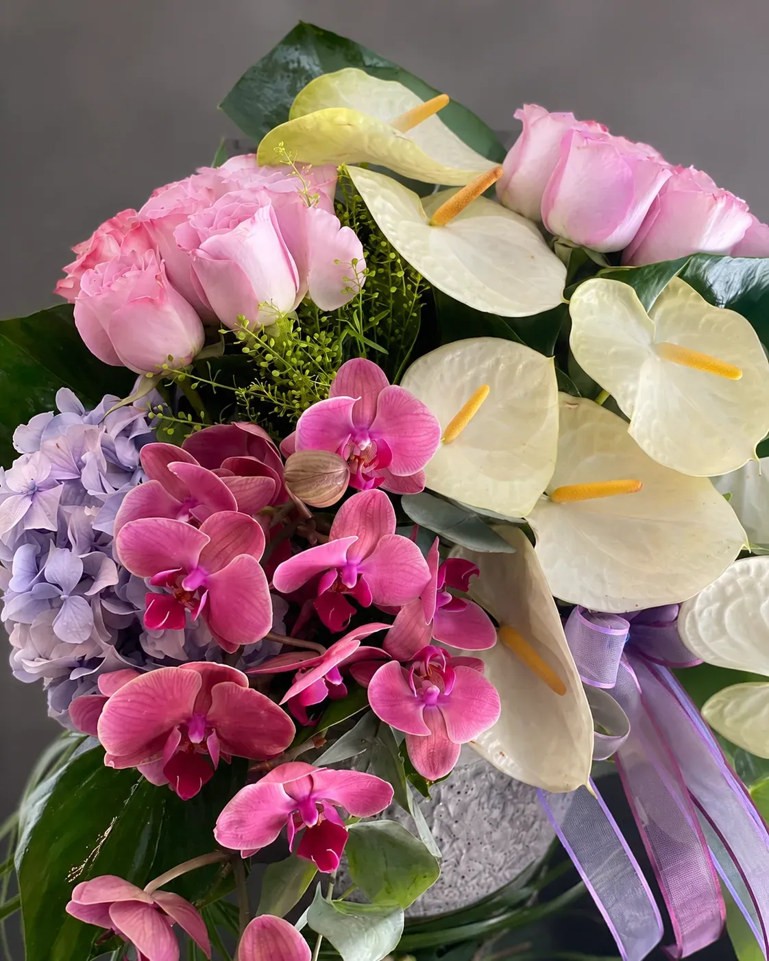 Beyaz anthurium, pembe orkide, pembe gül ve lila ortanca ile hazırlanmış gri vazoda tropikal çiçek aranjmanı